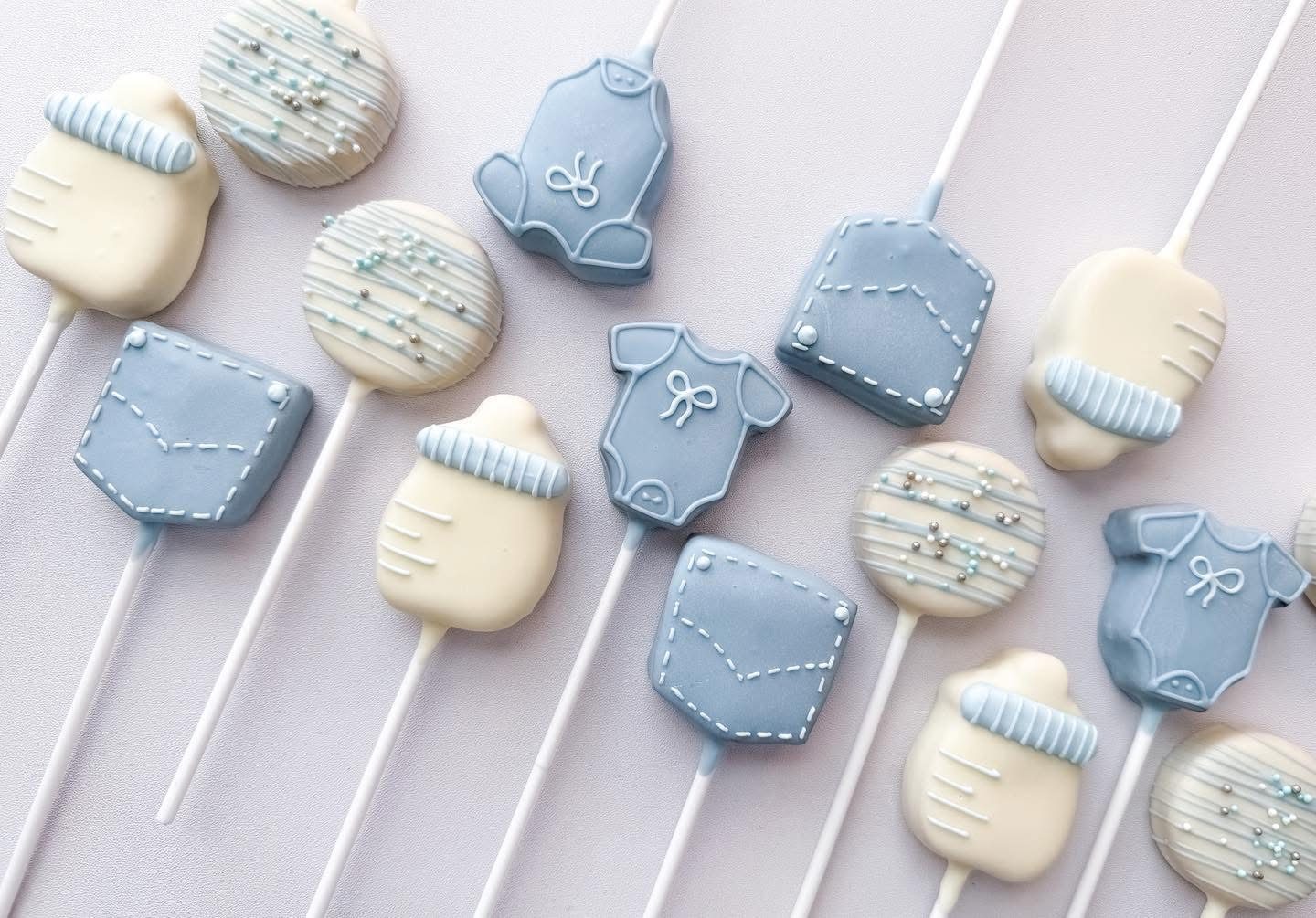 Blue baby shower cake pops with denim onesies, bottles, and pockets in white chocolate and soft blue with sprinkles