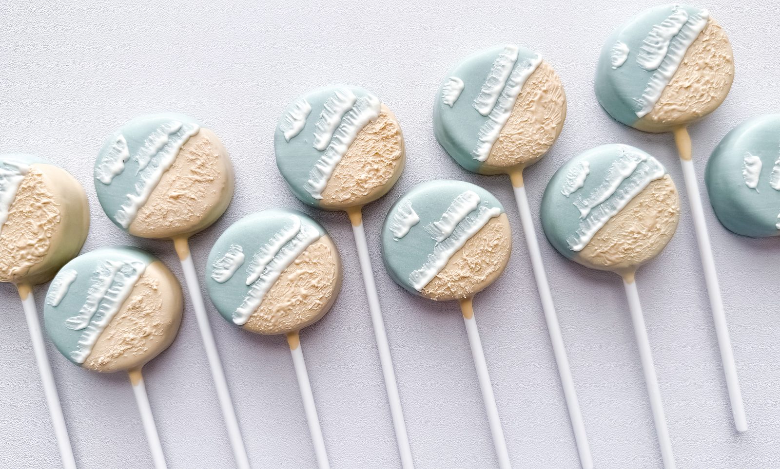 Coastal-themed cake pops half-dipped in soft blue and sandy cream with white wave drizzle