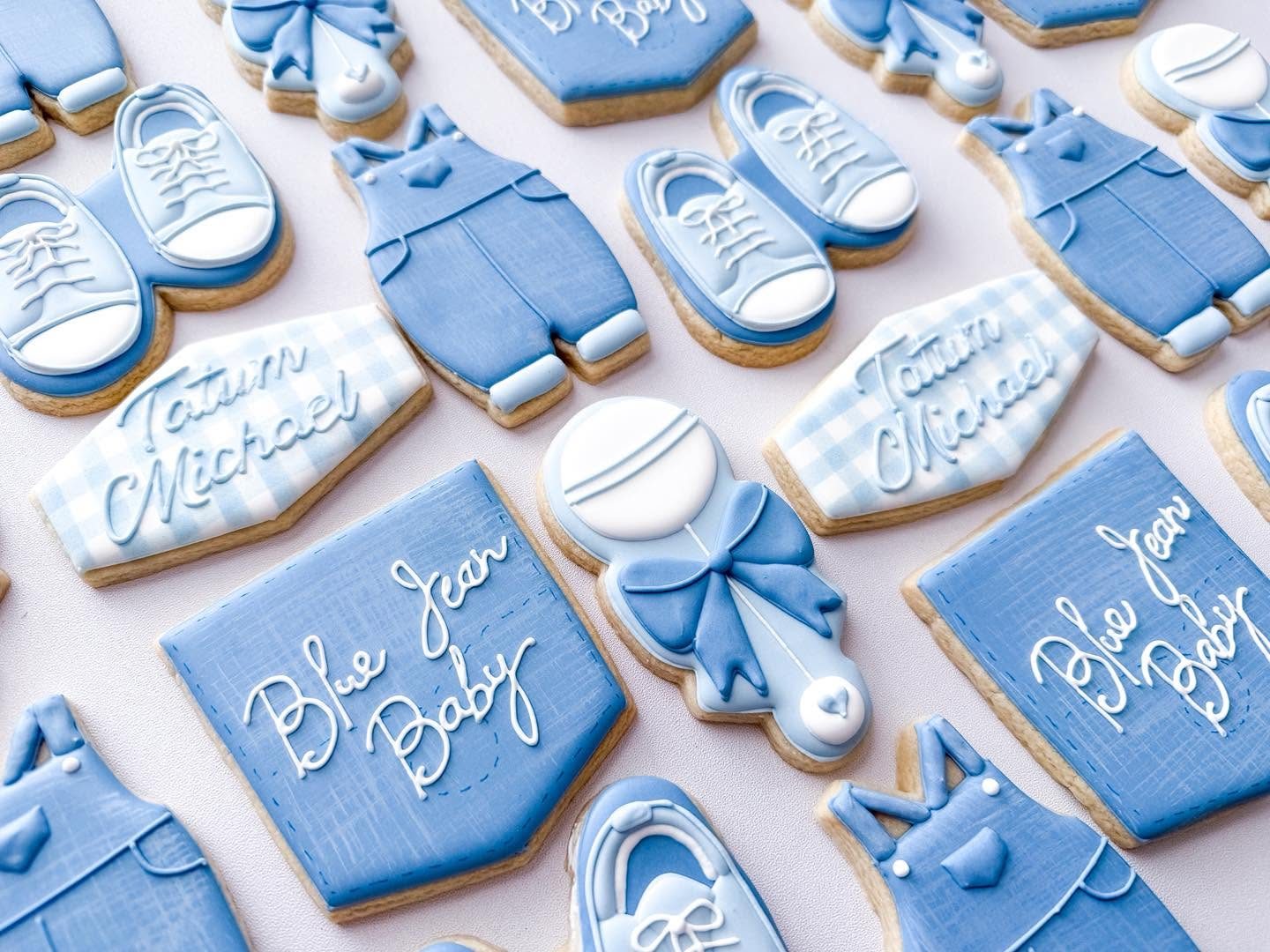 Blue Jean Baby denim-themed baby shower cookies with overalls, baby sneakers, gingham name plaques, rattles, and script lettering in shades of blue