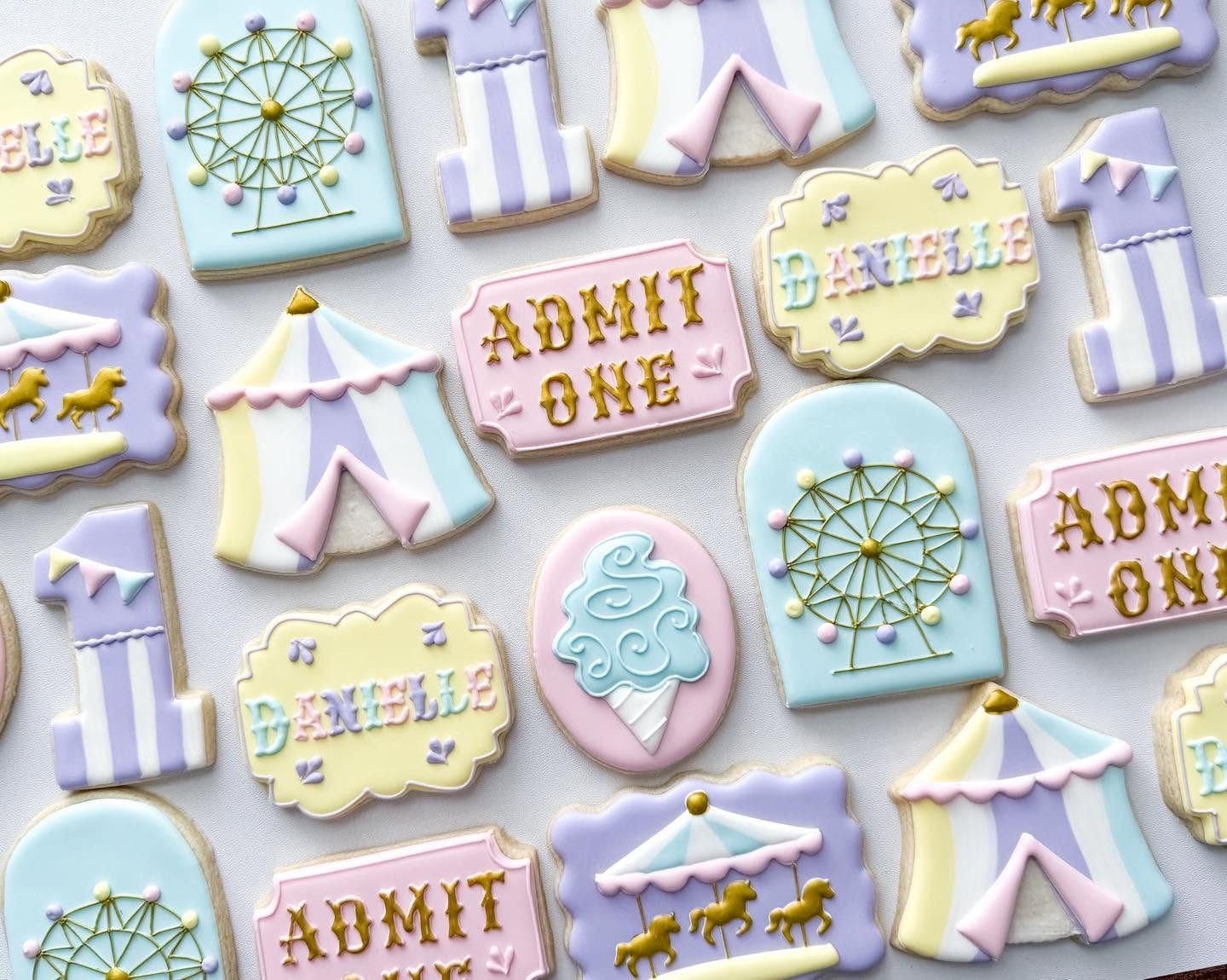 Pastel carnival first birthday cookies with Ferris wheels, circus tents, carousels, cotton candy, Admit One tickets, and name plaques in lavender, pink, yellow, and blue