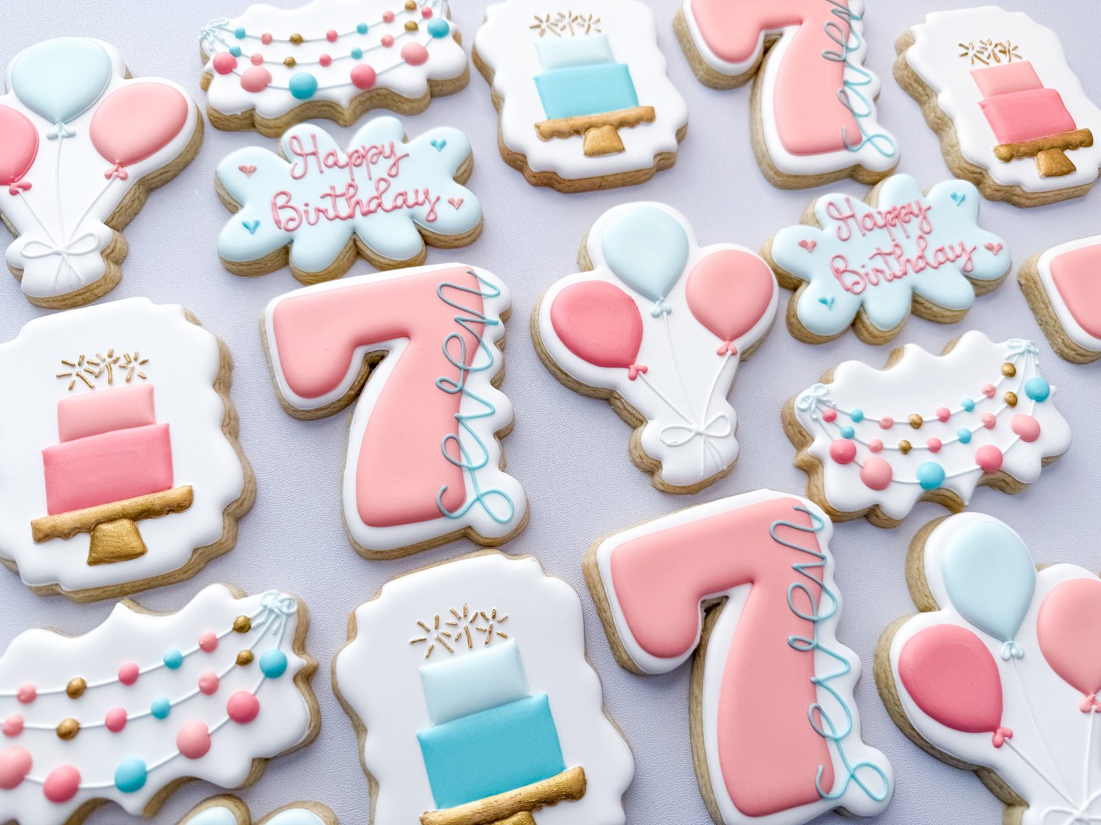 Seventh birthday cookies in coral and blue with balloons, tiered cakes, and party garland